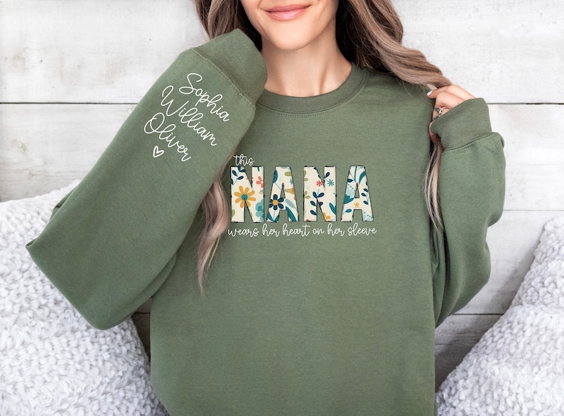 Op de afbeelding: Een groen sweatshirt met de tekst "This Nana wears her heart on her sleeve" en een bloemenontwerp. De mouw heeft de namen "Sophia, William, Oliver" erop geschreven.