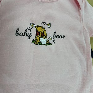 May include: A light pink baby onesie with a Winnie the Pooh embroidery. The embroidery features Winnie the Pooh with the words "baby bear" in a whimsical font. The onesie is made of soft fabric.