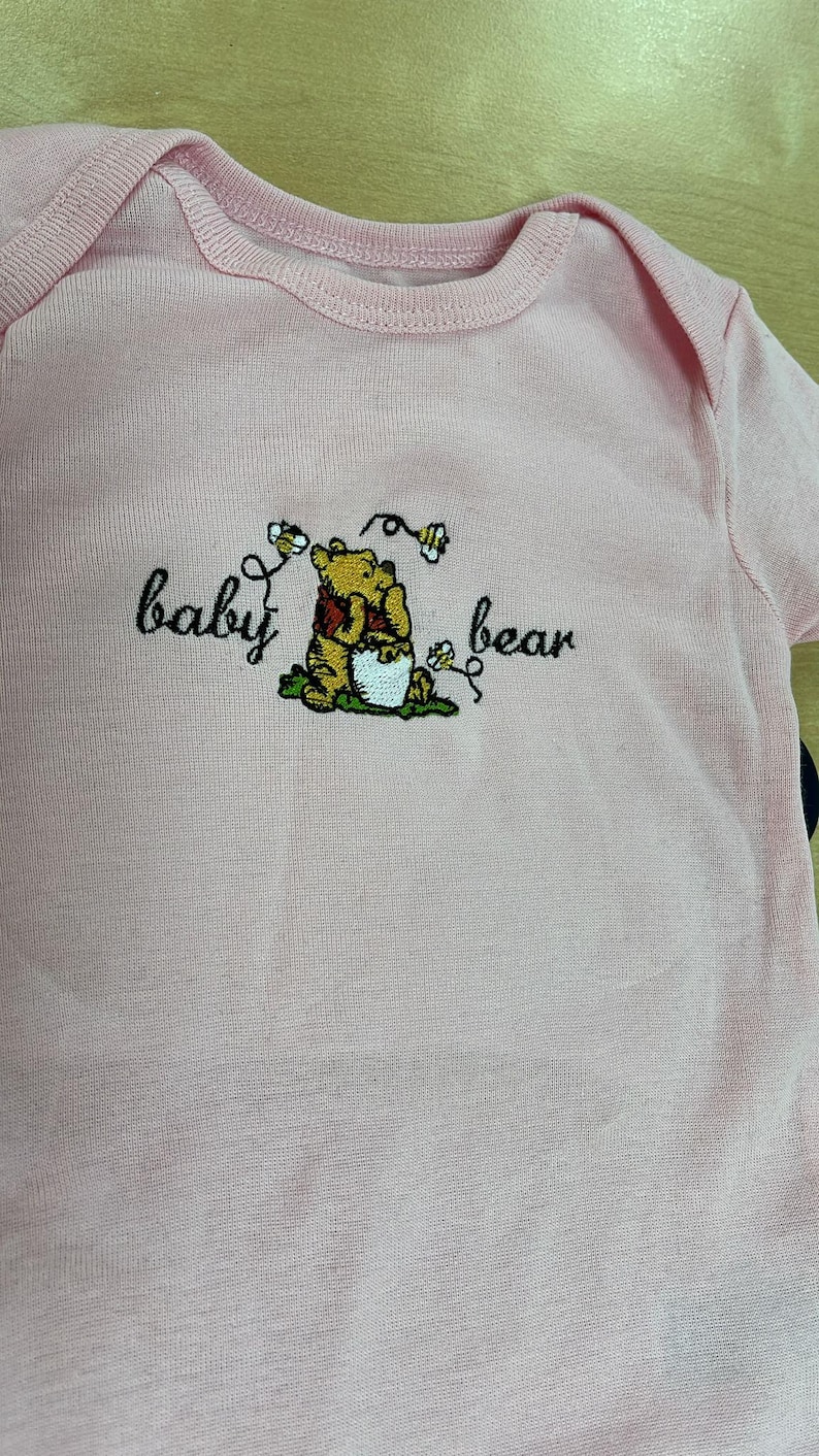 May include: A light pink baby onesie with a Winnie the Pooh embroidery. The embroidery features Winnie the Pooh with the words "baby bear" in a whimsical font. The onesie is made of soft fabric.