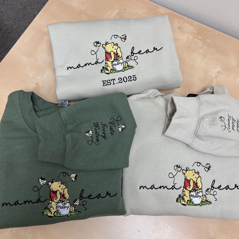 May include: Three sweatshirts in shades of green and beige. Each sweatshirt features an embroidered Winnie the Pooh design with the words "mama bear" and "EST. 2025". One sweatshirt has names embroidered on the sleeve.