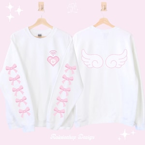 Puede incluir: Sudadera blanca de manga larga con lazos rosas en las mangas y un corazón rosa pixelado con un símbolo de wifi y un botón de encendido en la parte delantera. La parte posterior de la sudadera tiene alas de ángel rosas.