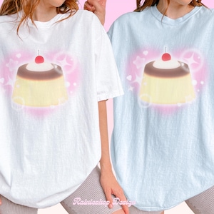 May include: Two short-sleeved crew neck t-shirts, one white and one light blue, each featuring a central graphic of a custard pudding topped with a red cherry. The design is surrounded by a soft pink airbrushed heart, white stars, and sparkles.