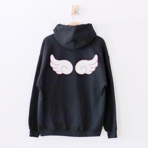 May include: Black hooded sweatshirt with a white angel wing design on the back. The wings have a pink outline.