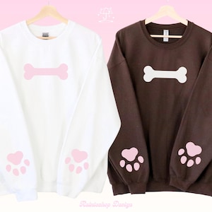 Könnte beinhalten: Zwei Sweatshirts, eines weiß und eines braun, jeweils mit einem Knochenmotiv auf der Brust und Pfotenabdrücken auf den Ärmeln. Das weiße Sweatshirt hat rosa Grafiken, das braune hat weiße Grafiken.