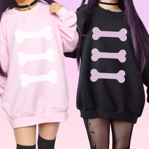 Pode incluir: Uma camisola de manga comprida cor-de-rosa claro com três gráficos de ossos brancos e uma camisola preta com três gráficos de ossos cor-de-rosa claro. Ambas as peças apresentam gola redonda, punhos canelados e um corte largo.