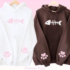 Op de afbeelding: Twee hoodies, één witte en één bruine, elk met een roze visgraatmotief op de voorkant. Roze pootafdrukken staan op de mouwen. De sweatshirts hangen aan een houten hanger.