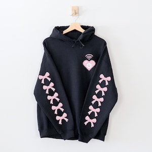 May include: Black hoodie with a pink pixelated heart with a wifi symbol and power button on the front. The sleeves have pink pixelated bows.