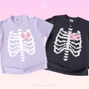 以下が含まれることがあります： ライトパープルとダークチャコールグレーの半袖クルーネックTシャツ2枚。各シャツの前面には、白いピクセル化された胸郭のグラフィックが描かれており、中央には小さな十字架のシンボルが入ったピンクのピクセル化されたハートの形が配置されています。