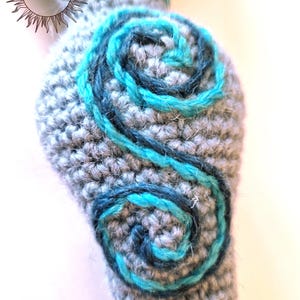 May include: A gray crocheted object featuring two dark blue and bright turquoise yarn spirals embroidered on its surface. The text MERRY MANDRAKE is visible in the upper left corner within a sunburst graphic design on the crocheted fabric.