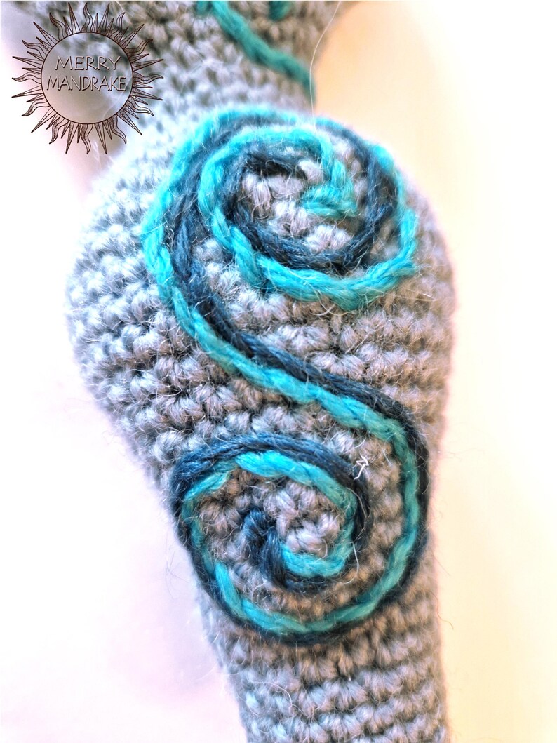 May include: A gray crocheted object featuring two dark blue and bright turquoise yarn spirals embroidered on its surface. The text MERRY MANDRAKE is visible in the upper left corner within a sunburst graphic design on the crocheted fabric.