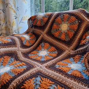 May include: A crocheted throw blanket with a brown, orange, and blue geometric pattern. The blanket is made of soft yarn and has a textured surface.