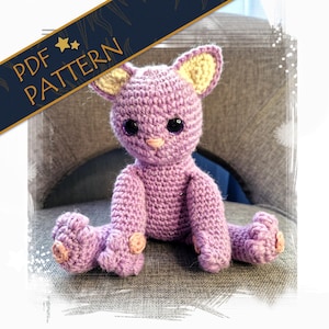 May include: A handmade crochet cat in shades of purple, with yellow-lined ears and a peach-colored nose and paw pads. The cat is sitting with its legs outstretched. The image includes text that reads "PDF PATTERN".