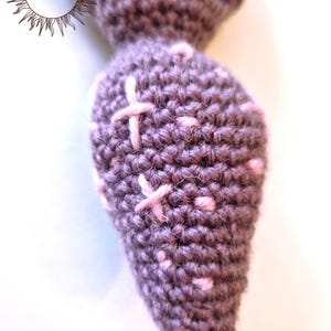 May include: A brown crocheted mandrake root with a tapered shape, featuring light pink embroidered cross-stitch details and small pink dots scattered across the surface. The text MERRY MANDRAKE is visible in the upper left corner of the image.