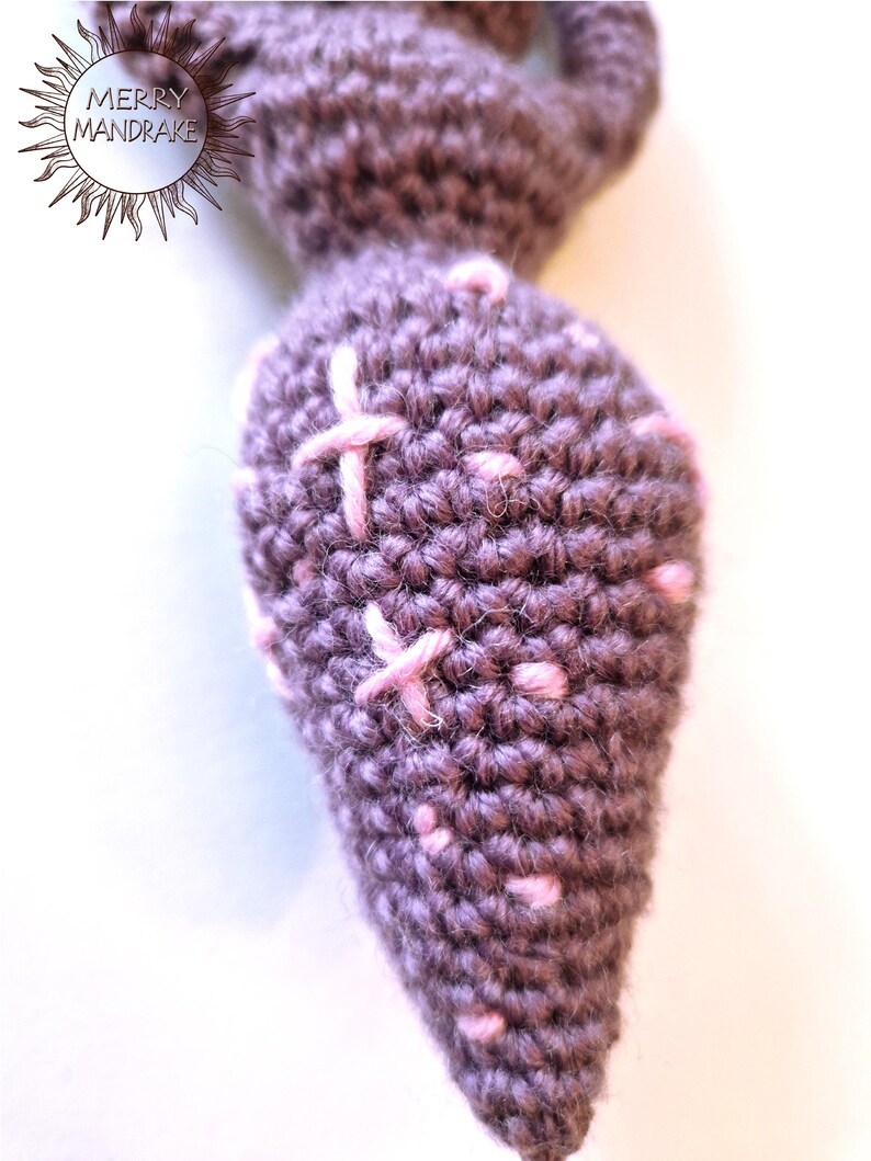 May include: A brown crocheted mandrake root with a tapered shape, featuring light pink embroidered cross-stitch details and small pink dots scattered across the surface. The text MERRY MANDRAKE is visible in the upper left corner of the image.