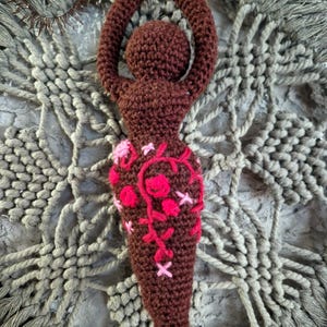 May include: A dark brown crocheted figurine with a rounded head and arms raised in a circular shape above the head. The lower body features embroidered floral patterns in shades of bright pink and light pink, with a small logo in the top corner.