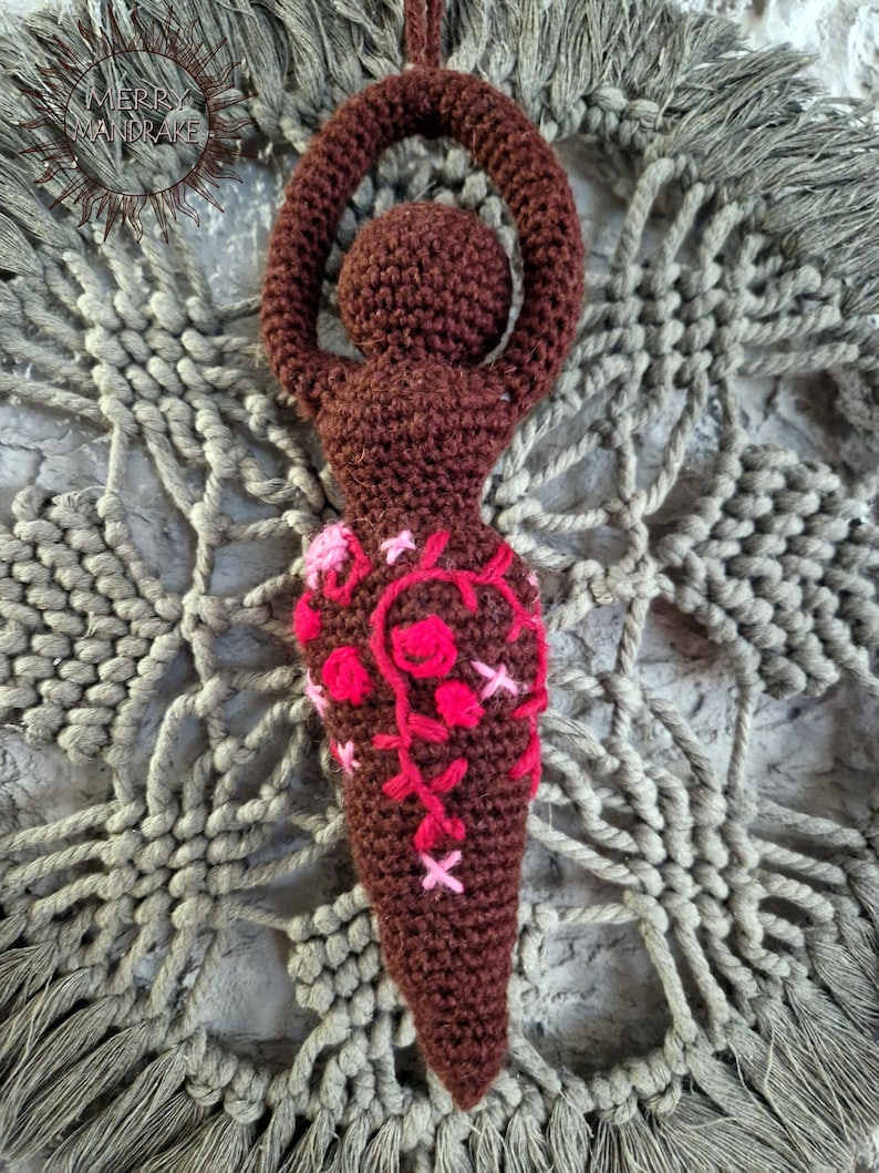 May include: A dark brown crocheted figurine with a rounded head and arms raised in a circular shape above the head. The lower body features embroidered floral patterns in shades of bright pink and light pink, with a small logo in the top corner.