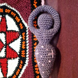 May include: A purple crocheted goddess figurine with a circular head and arms raised in a loop, featuring light pink embroidered cross stitches on the torso. The figure hangs by a light pink yarn loop next to a square tapestry with the text MERRY MANDRAKE.