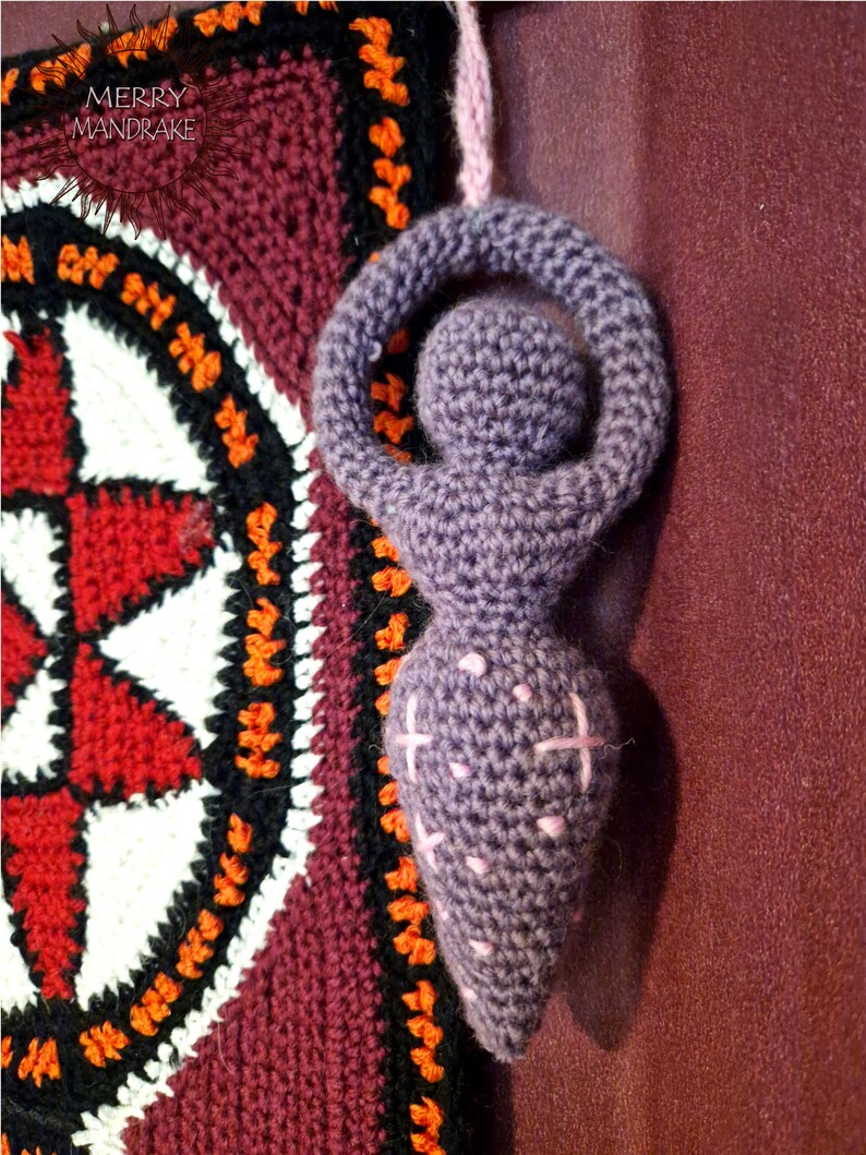May include: A purple crocheted goddess figurine with a circular head and arms raised in a loop, featuring light pink embroidered cross stitches on the torso. The figure hangs by a light pink yarn loop next to a square tapestry with the text MERRY MANDRAKE.