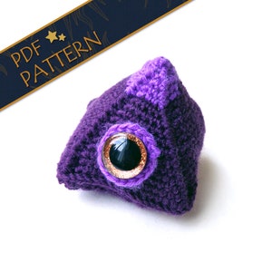 May include: Purple crocheted amigurumi toy with a large black eye and a purple patch on the top. The toy is shaped like a triangle.