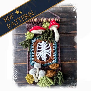 May include: A handmade crochet wall hanging featuring a whimsical design. The piece includes red and white mushroom accents, green foliage, and a central square with a white, stylized design. The text "PDF PATTERN" is displayed on a banner.