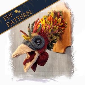 Op de afbeelding: Een gehaakte haan kop met een kleurrijke, getextureerde kam, een grote open snavel en een grijs oog. De haan kop is gemonteerd op een houten plaat.