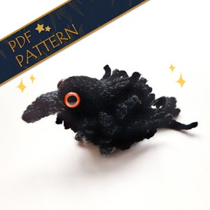 Könnte beinhalten: Ein schwarzer gehäkelter Rabe mit orangefarbenen Augen. Der Rabe ist ein kleines Stofftier, perfekt für ein Kinderspielzeug oder ein Sammlerstück.