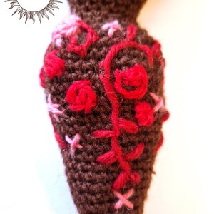May include: A brown crocheted mandrake root featuring embroidered red and pink floral patterns. The surface is textured with yarn stitches, and the upper portion includes a circular logo with the text MERRY MANDRAKE in black ink.