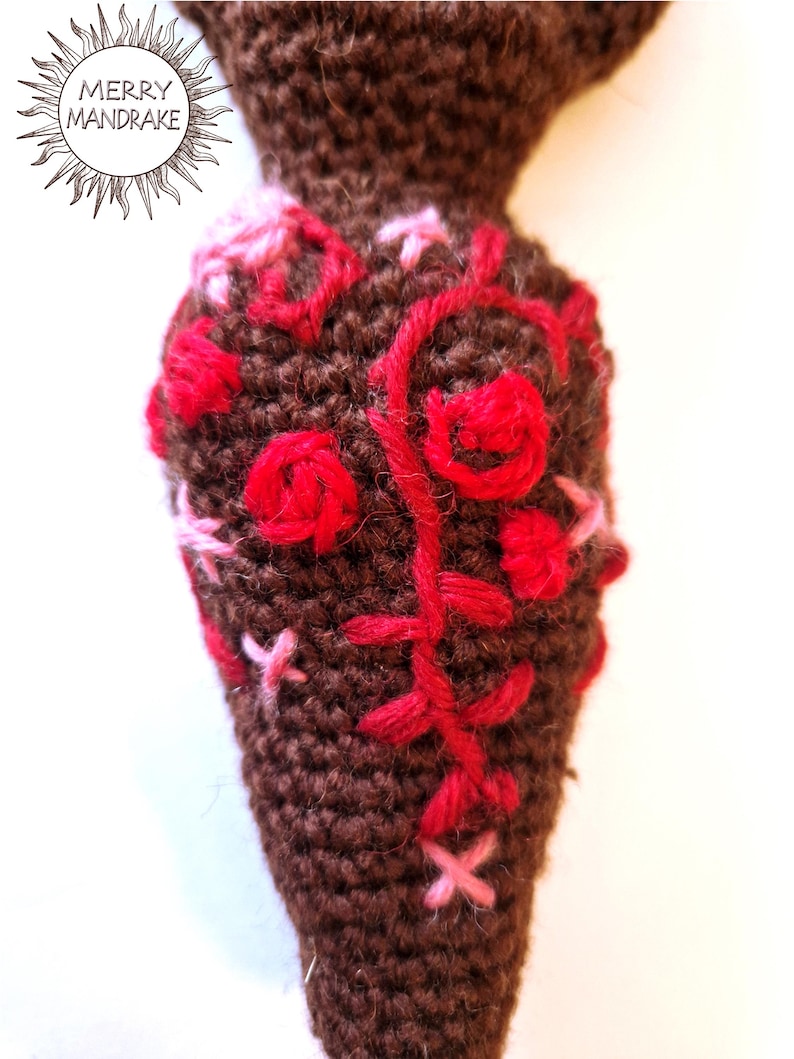 May include: A brown crocheted mandrake root featuring embroidered red and pink floral patterns. The surface is textured with yarn stitches, and the upper portion includes a circular logo with the text MERRY MANDRAKE in black ink.