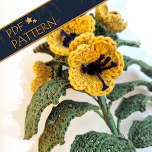 Puede incluir: Primer plano de un arreglo floral tejido a ganchillo con flores amarillas, centros azul oscuro y hojas verdes. El texto "PDF PATTERN" está en una pancarta azul marino. Las flores y hojas hechas a mano tienen una textura.