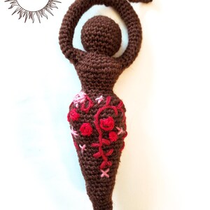 May include: A dark brown crocheted figurine with a rounded head, raised arms forming a loop, and a tapered lower body. The midsection features decorative embroidery in shades of red and pink thread. A small loop extends from the top of the head.