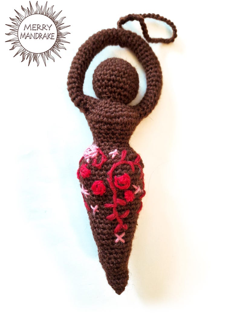 May include: A dark brown crocheted figurine with a rounded head, raised arms forming a loop, and a tapered lower body. The midsection features decorative embroidery in shades of red and pink thread. A small loop extends from the top of the head.