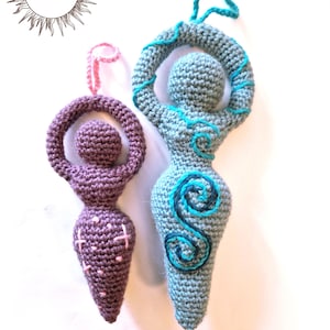 May include: Two crochet goddess figurines with hanging loops. The smaller one is purple with pink stitched accents. The larger one is light blue with dark blue and teal spiral embroidery. The text MERRY MANDRAKE appears in a sun-shaped logo.