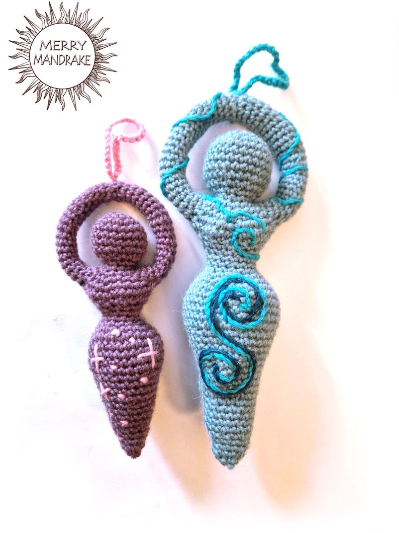 May include: Two crochet goddess figurines with hanging loops. The smaller one is purple with pink stitched accents. The larger one is light blue with dark blue and teal spiral embroidery. The text MERRY MANDRAKE appears in a sun-shaped logo.