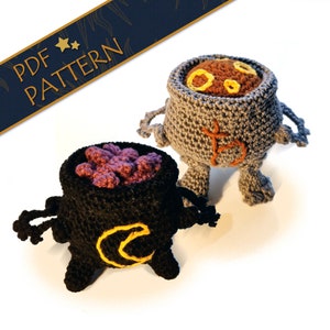 May include: Two crocheted cauldron amigurumi toys. One is grey with orange and yellow details and the other is black with purple and yellow details. Both have small legs and arms.