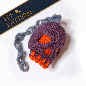 Könnte beinhalten: Ein gehäkelter Totenkopf-Anhänger mit einem violetten Schädel und orangefarbenen Details. Der Schädel ist an einer grauen Kette befestigt.
