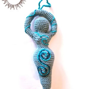May include: A grey crocheted figurine with a rounded head, raised arms forming a circle, and a tapered body. The figure features embroidered light blue and dark blue spiral patterns on the chest and abdomen, with a small loop at the top for hanging.