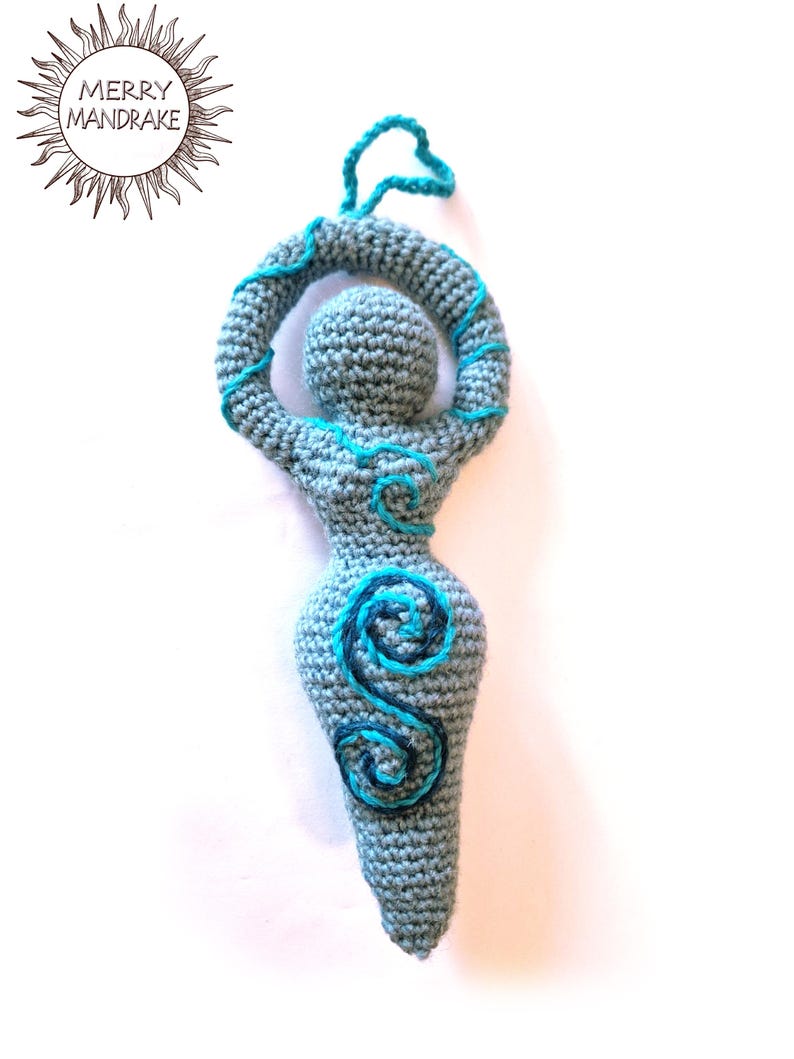 May include: A grey crocheted figurine with a rounded head, raised arms forming a circle, and a tapered body. The figure features embroidered light blue and dark blue spiral patterns on the chest and abdomen, with a small loop at the top for hanging.