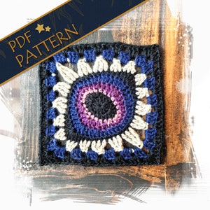 Puede incluir: Un cuadrado de crochet con un esquema de color negro, azul, morado y blanco. El cuadrado tiene un diseño circular en el centro con un círculo negro rodeado de anillos morados, azules y blancos. El cuadrado está hecho con un punto texturizado y tiene un borde de flecos.