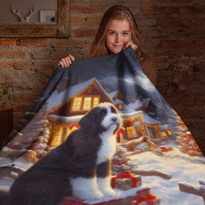 May include: A rectangular throw blanket featuring a printed scene of a black and white dog sitting in the snow in front of a cabin decorated for the holidays. The cabin has glowing yellow windows and the ground is covered in snow with gift boxes.