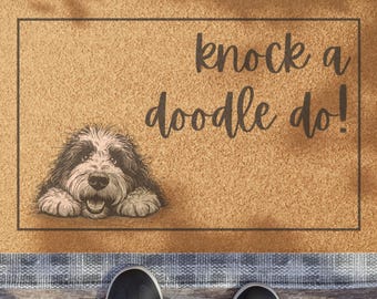 Könnte beinhalten: Eine rechteckige Fußmatte mit brauner Mitte und grauem Karomuster. Die Matte zeigt eine Schwarz-Weiß-Illustration eines Hundes und den Satz "knock a doodle do!" in schwarzer Schrift.