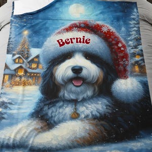 May include: A plush, blue blanket featuring a Bernese Mountain Dog wearing a Santa hat with the name "Bernie" on it. The dog is set against a winter scene with a full moon, snow, and a Christmas tree.