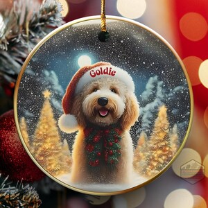 May include: A round Christmas ornament featuring a smiling Goldendoodle wearing a Santa hat with the name "Goldie" and a festive scarf. The background shows a snowy forest scene with Christmas trees and a full moon. The ornament has a gold trim.