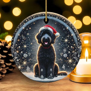 May include: A round Christmas ornament featuring a black dog wearing a Santa hat and a red collar, set against a snowy, starry background. The ornament hangs from a gold string. A lit candle and pinecones are in the foreground.