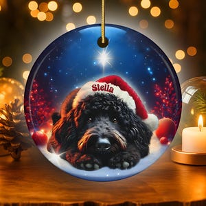 May include: Round Christmas ornament featuring a black dog wearing a Santa hat with the name "Stella". The ornament has a blue background with a star and snow effect. A gold string is attached for hanging.