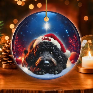 May include: Round Christmas ornament featuring a black dog wearing a Santa hat, with the name "Stella" on the hat. The ornament has a blue background with a star and snow. A gold string is attached for hanging.