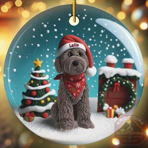 May include: A round Christmas ornament featuring a brown dog wearing a Santa hat with the name "Latte" on it and a red bandana. The ornament has a Christmas tree, a fireplace, and snow. The background is blurred with Christmas lights.