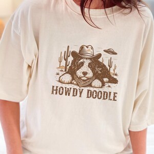Może przedstawiać: Kremowa koszulka z brązową grafiką psa w kapeluszu kowbojskim i chuście, z kaktusami i UFO. Pod spodem znajduje się napis "HOWDY DOODLE". Koszulka ma kr&oacute;tkie rękawy i luźny kr&oacute;j.