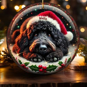 May include: A round Christmas ornament featuring a black dog wearing a Santa hat. The dog's name, "Stella", is printed on the hat. The ornament is decorated with holly and berries, and has a gold string for hanging.