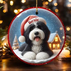 May include: Round Christmas ornament featuring a black and white dog wearing a Santa hat with the name "Bernie". The ornament has a red border and a winter scene with a house and snow. A gold string is attached for hanging.