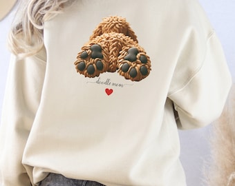 May include: Beige sweatshirt featuring a brown doodle dog paw print design with the words "doodle mom" and a red heart. The sweatshirt is a neutral color and has a crew neck.